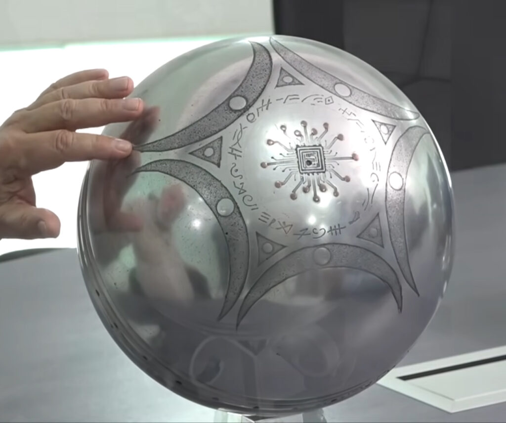 A esfera metálica encontrada em Buga, exibindo glifos (grafemas) e marcas semelhantes a circuitos eletrônicos em sua superfície.